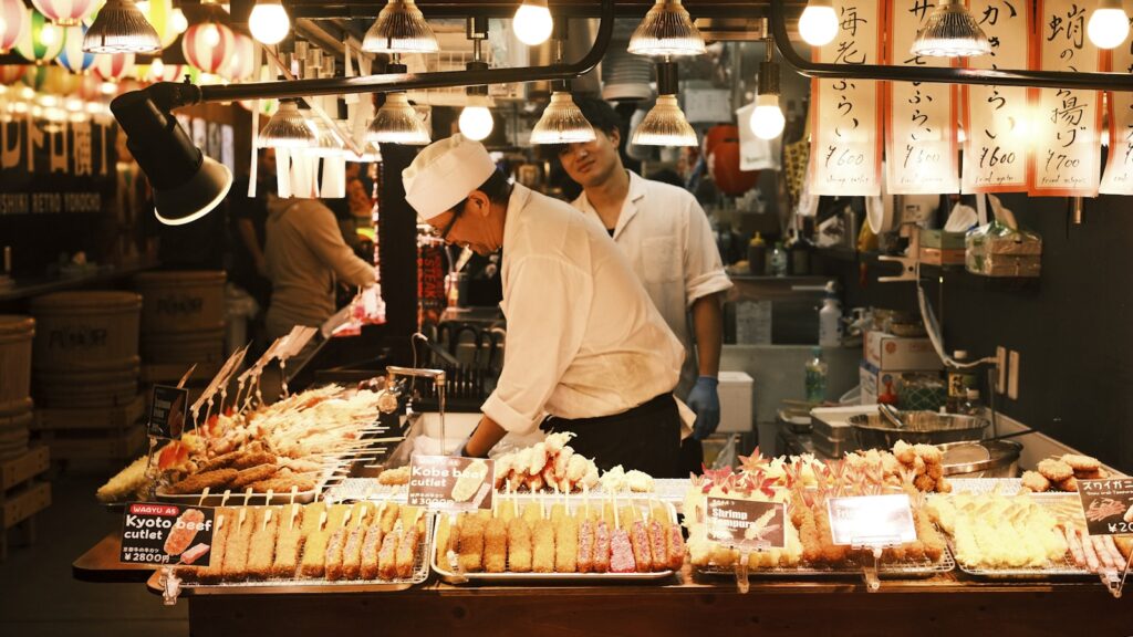 市場の屋台に並ぶ串料理。賑わう食市場（錦市場とみられる）の情景。フードツーリズムのイメージ