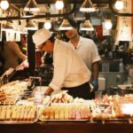 市場の屋台に並ぶ串料理。賑わう食市場（錦市場とみられる）の情景。フードツーリズムのイメージ