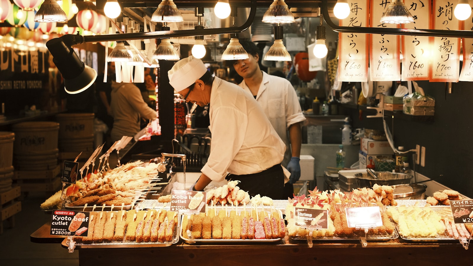市場の屋台に並ぶ串料理。賑わう食市場（錦市場とみられる）の情景。フードツーリズムのイメージ