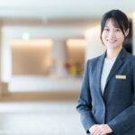 明るいホテルのロビーに立つ名札を付けたスタッフの女性。観光人材・ホスピタリティのイメージ