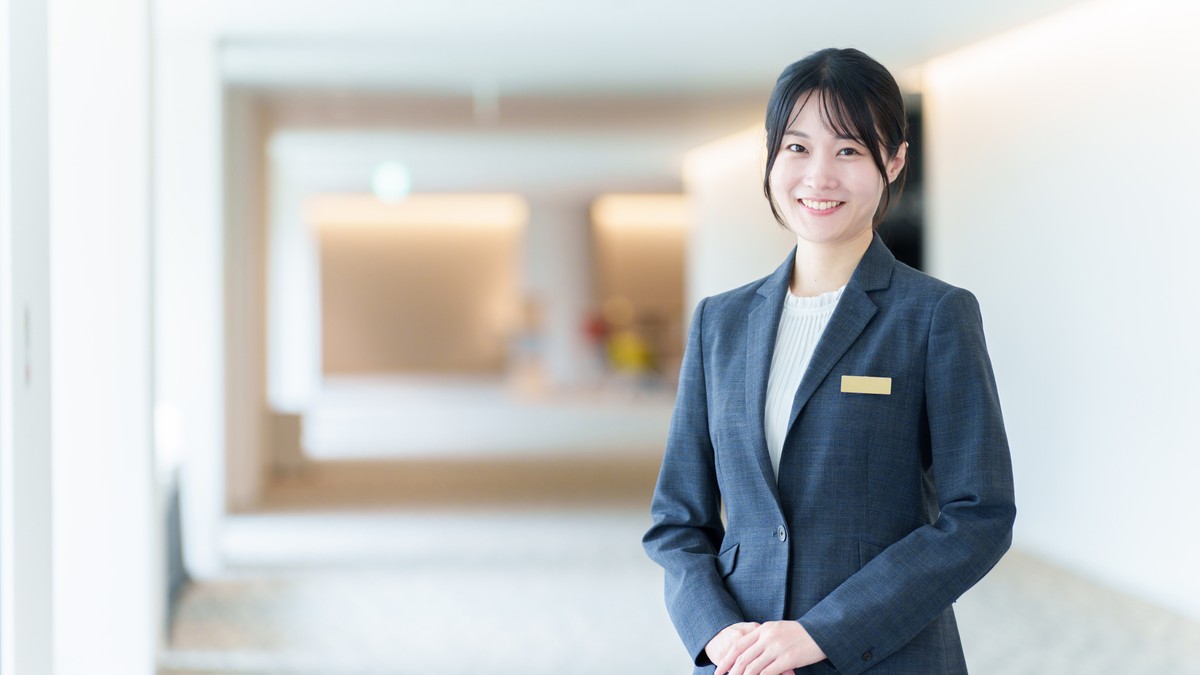 明るいホテルのロビーに立つ名札を付けたスタッフの女性。観光人材・ホスピタリティのイメージ