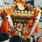 御大典の幟を掲げて担がれる金色の神輿と担ぎ手たち。祭りでにぎわう地域の情景。地方創生・DMOのイメージ