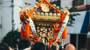 御大典の幟を掲げて担がれる金色の神輿と担ぎ手たち。祭りでにぎわう地域の情景。地方創生・DMOのイメージ