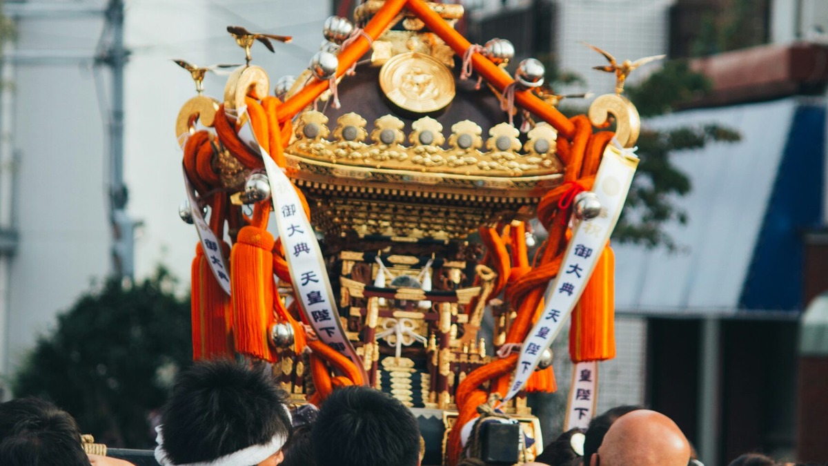 御大典の幟を掲げて担がれる金色の神輿と担ぎ手たち。祭りでにぎわう地域の情景。地方創生・DMOのイメージ