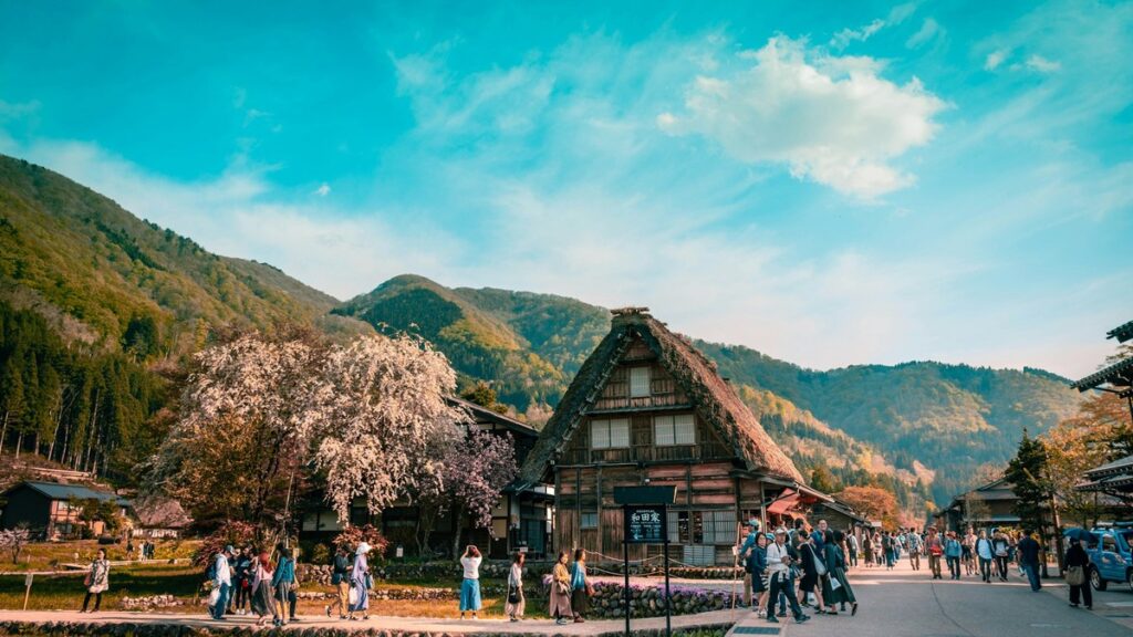 白川郷・和田家周辺を桜のもと歩く観光客と背後の山並み。持続可能な観光（サステナブルツーリズム）のイメージ