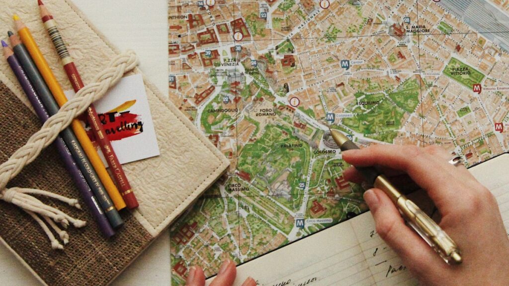 地図の上に置かれた眼鏡と色鉛筆、旅程を書き込む手。旅行業界の全体像ハブのヒーロー画像。
