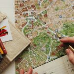 地図の上に置かれた眼鏡と色鉛筆、旅程を書き込む手。旅行業界の全体像ハブのヒーロー画像。