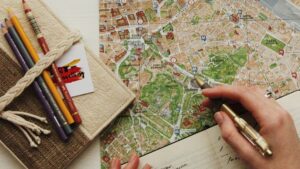 地図の上に置かれた眼鏡と色鉛筆、旅程を書き込む手。旅行業界の全体像ハブのヒーロー画像。