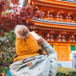 紅葉した木々と社殿を背に立つ、ニット帽をかぶった旅行者の後ろ姿