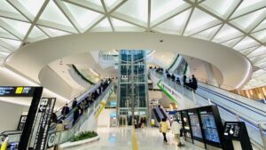 空港ターミナルの吹き抜け。三角格子の天窓から光が差し、エスカレーターを上り下りする利用者と到着案内板が見える
