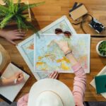 机に広げた世界地図を指し示す手元。帽子やカメラ、コーヒーが並ぶ
