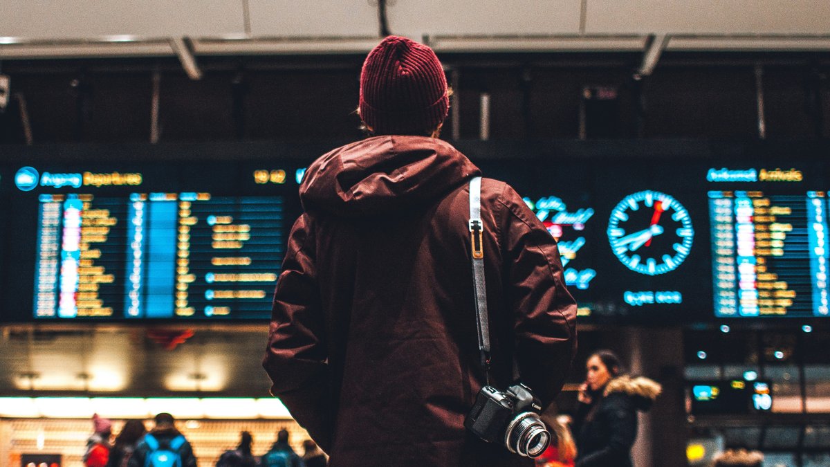 空港の出発案内板を見上げる旅行者。航空券流通の入り口となる運航情報が並ぶ