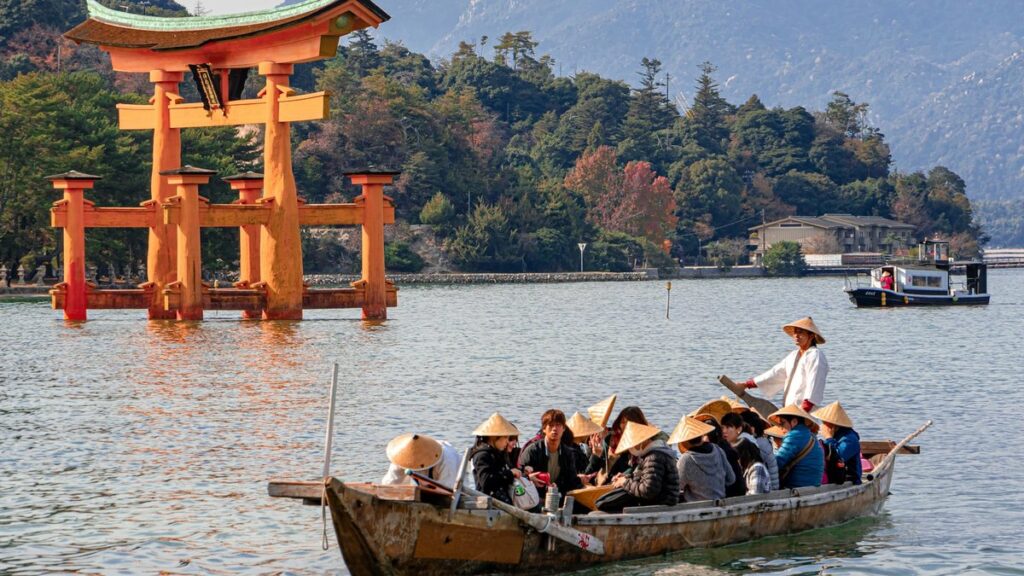 宮島・厳島神社の朱塗りの大鳥居の前を進む渡し船と、菅笠をかぶって乗る観光客。モニターツアーで観光地を体験するイメージ