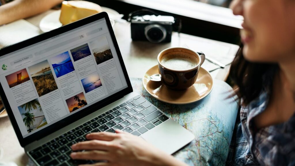 カフェでノートパソコンを開き、旅行先の写真を眺める人。オンラインツアーで旅の風景を画面越しに楽しむイメージ