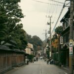 格子戸と瓦屋根の町家が並ぶ古い街道筋を、数人の歩行者が行き交う。電柱と電線が続き、突き当たりは紅葉した山につながる。