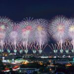 市街地の夜空に一斉に開く花火。手前に街明かりが広がる