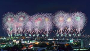 市街地の夜空に一斉に開く花火。手前に街明かりが広がる