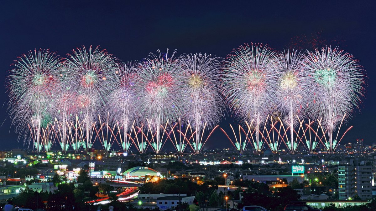 市街地の夜空に一斉に開く花火。手前に街明かりが広がる