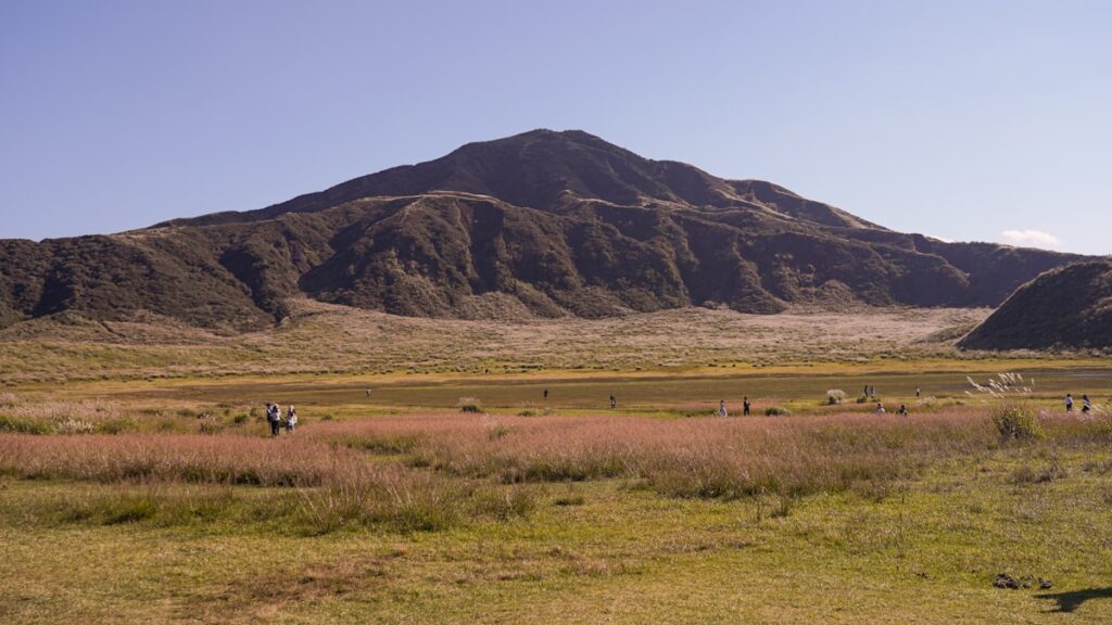 草原の向こうにそびえる阿蘇の山並み。手前のススキと草地を、点在する人影が歩いている