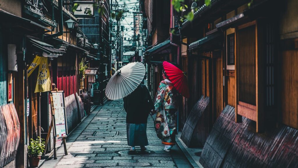 石畳の路地を和傘を差して歩く着物姿の2人。両側に町家が続く