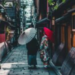 石畳の路地を和傘を差して歩く着物姿の2人。両側に町家が続く