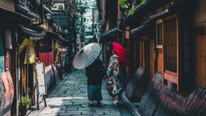 石畳の路地を和傘を差して歩く着物姿の2人。両側に町家が続く