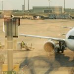 夕方の空港エプロン。ジェットブリッジに接続した白い旅客機と、手前を横切る作業車両、奥に管制塔が見える