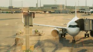 夕方の空港エプロン。ジェットブリッジに接続した白い旅客機と、手前を横切る作業車両、奥に管制塔が見える