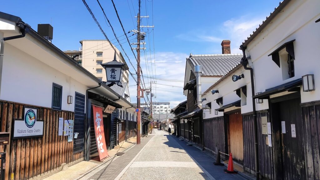 白壁と焼杉板の酒蔵が両側に並ぶ通り。左手に「黄桜」の看板行灯と黄桜カッパカントリーの案内板が見える