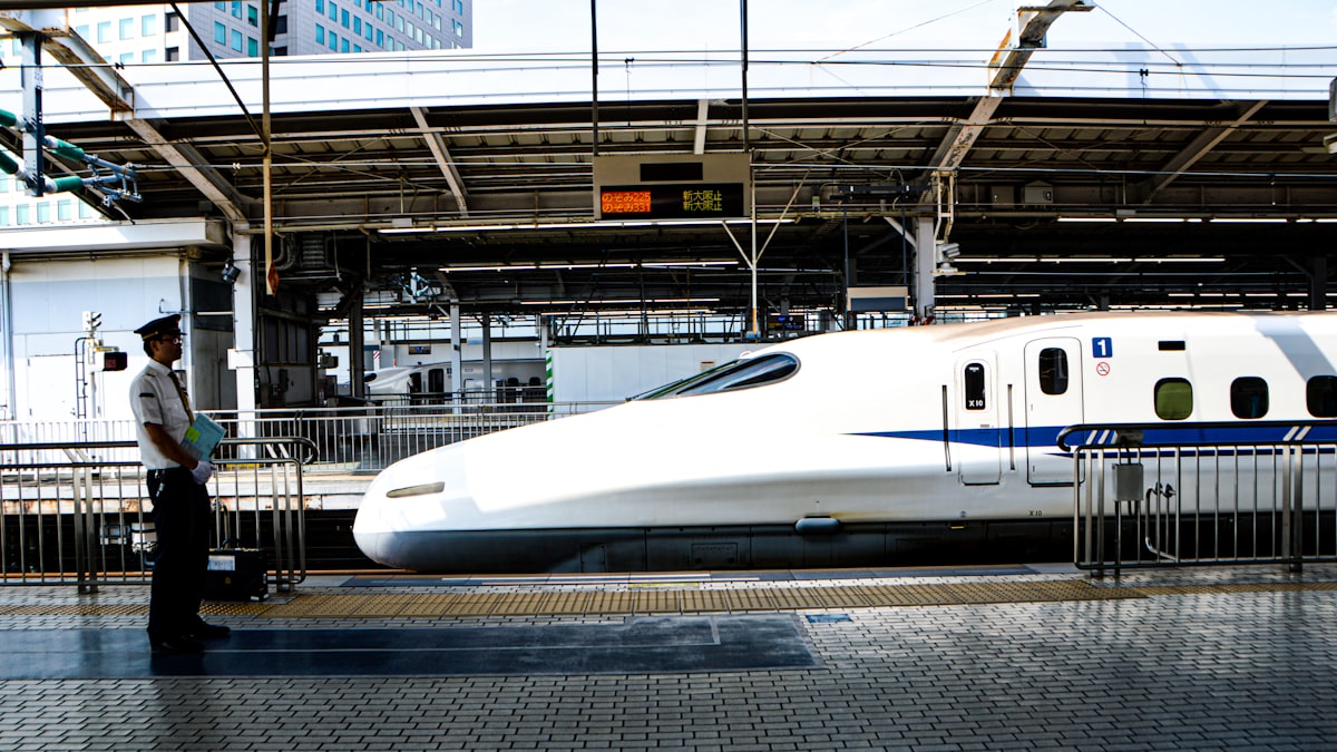 新幹線のホームに立つ駅係員と、停車中の白い新幹線。奥の発車案内には新大阪行きが表示されている