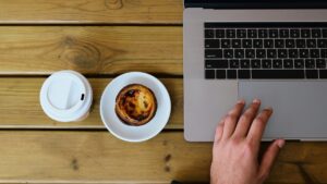 木のテーブルでノートパソコンを操作する手元と、テイクアウトのコーヒー、皿にのったエッグタルト