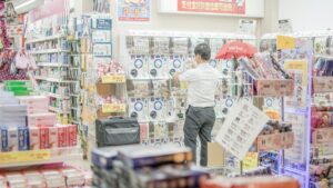 大阪の量販店の店内。ガチャガチャの機械が並ぶ売り場で男性客が商品を手に取り、奥の壁には中国の決済サービスが使えることを知らせる掲示が出ている