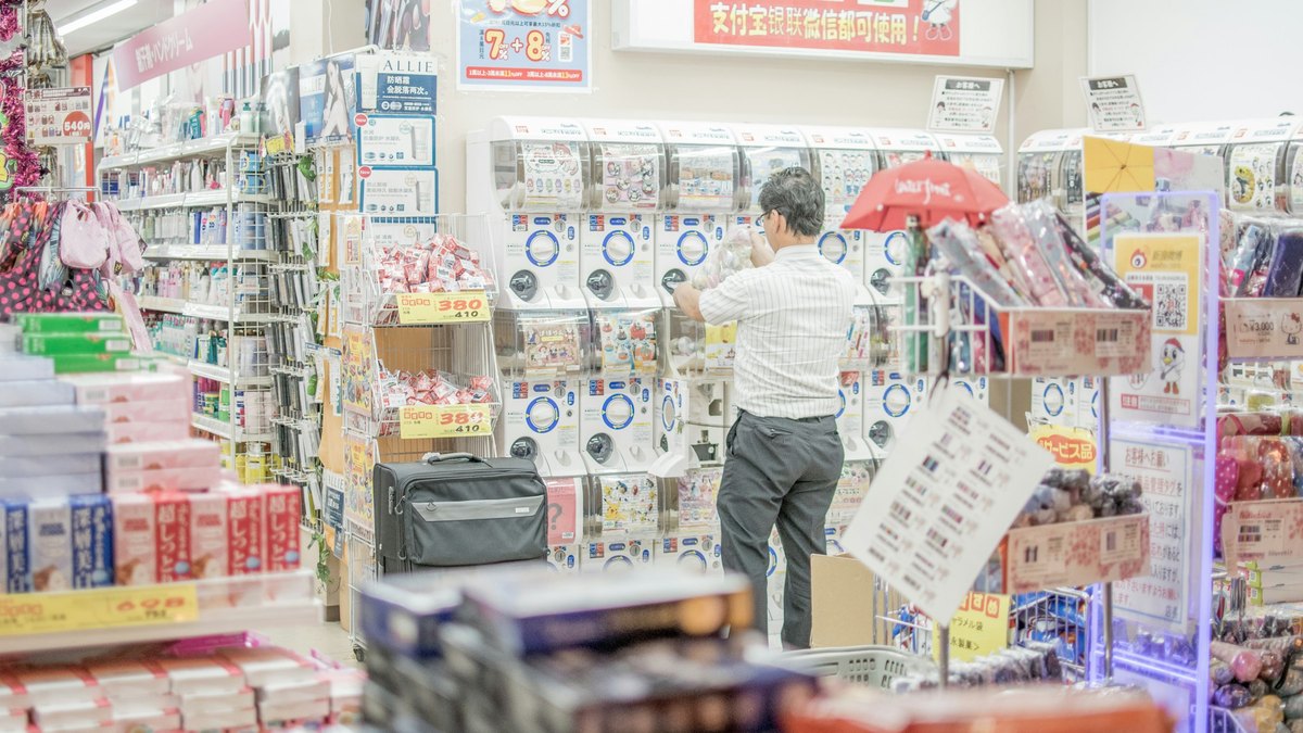 大阪の量販店の店内。ガチャガチャの機械が並ぶ売り場で男性客が商品を手に取り、奥の壁には中国の決済サービスが使えることを知らせる掲示が出ている