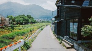 山を背にした地方の町で、川沿いの細い道を自転車の人が走り、右に黒い板張りの建物が建つ風景