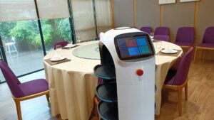 円卓のある飲食店の個室で、白い配膳ロボットが画面を見せて立っている様子