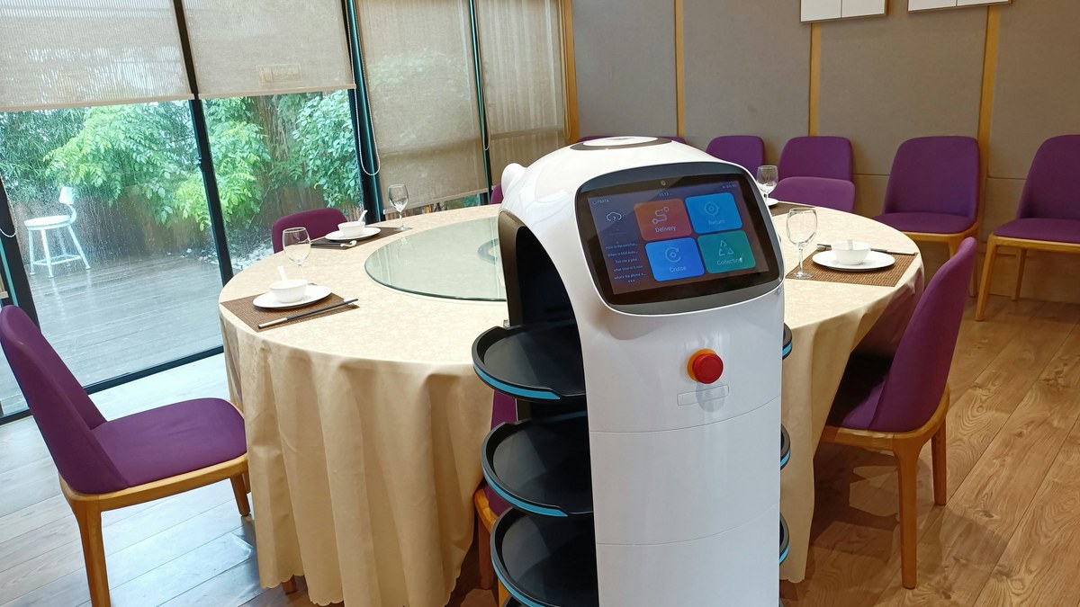 円卓のある飲食店の個室で、白い配膳ロボットが画面を見せて立っている様子