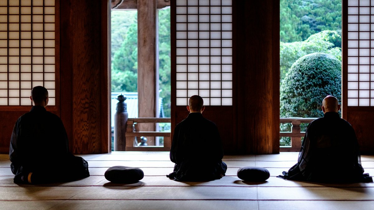 寺院の堂内で、庭に向かって坐禅を組む三人の後ろ姿