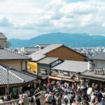 瓦屋根の商店が並ぶ坂道に集まる観光客。奥に山並みが見える
