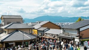 瓦屋根の商店が並ぶ坂道に集まる観光客。奥に山並みが見える