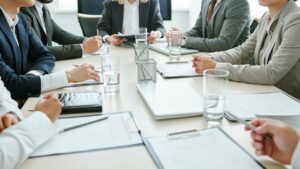 会議室の卓を囲み、書類を前にペンを手にしたスーツ姿の参加者たち